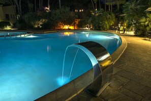 Ein Pool mit geschwungener Kante, umgeben von einer gepflasterten Fläche mit Stühlen und Tischen, und ein Gebäude mit beleuchteten Fenstern im Hintergrund.