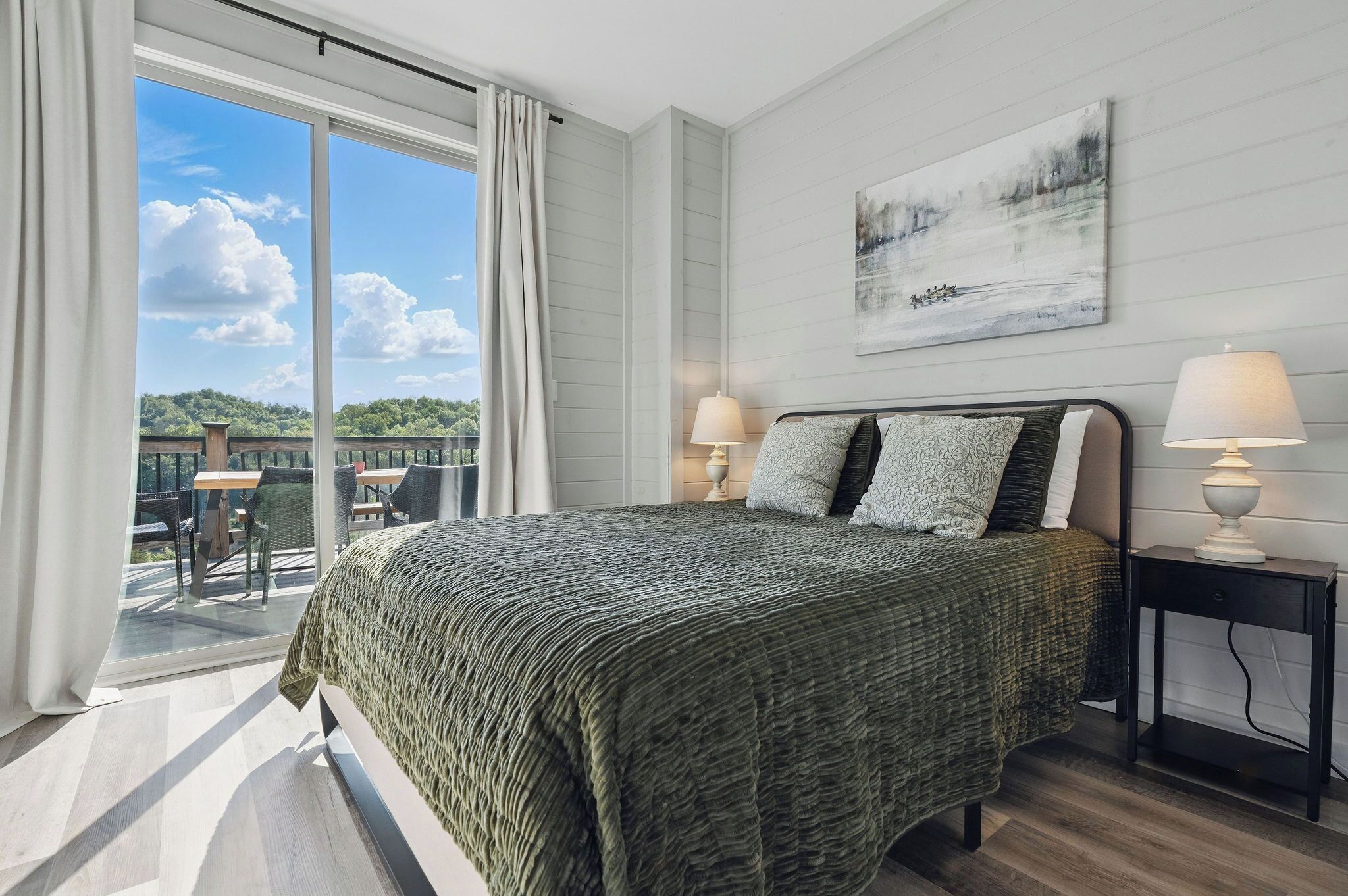 A hotel room with a large bed, two bedside lamps, a painting on the wall, and a balcony with outdoor furniture.