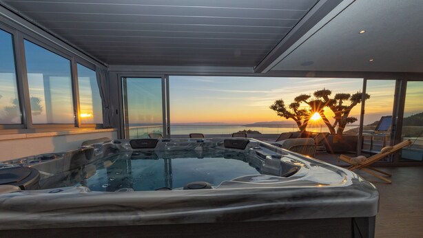 Un moderno jacuzzi con cerramiento de vidrio, ventanas amplias y vistas a la puesta de sol sobre el océano.