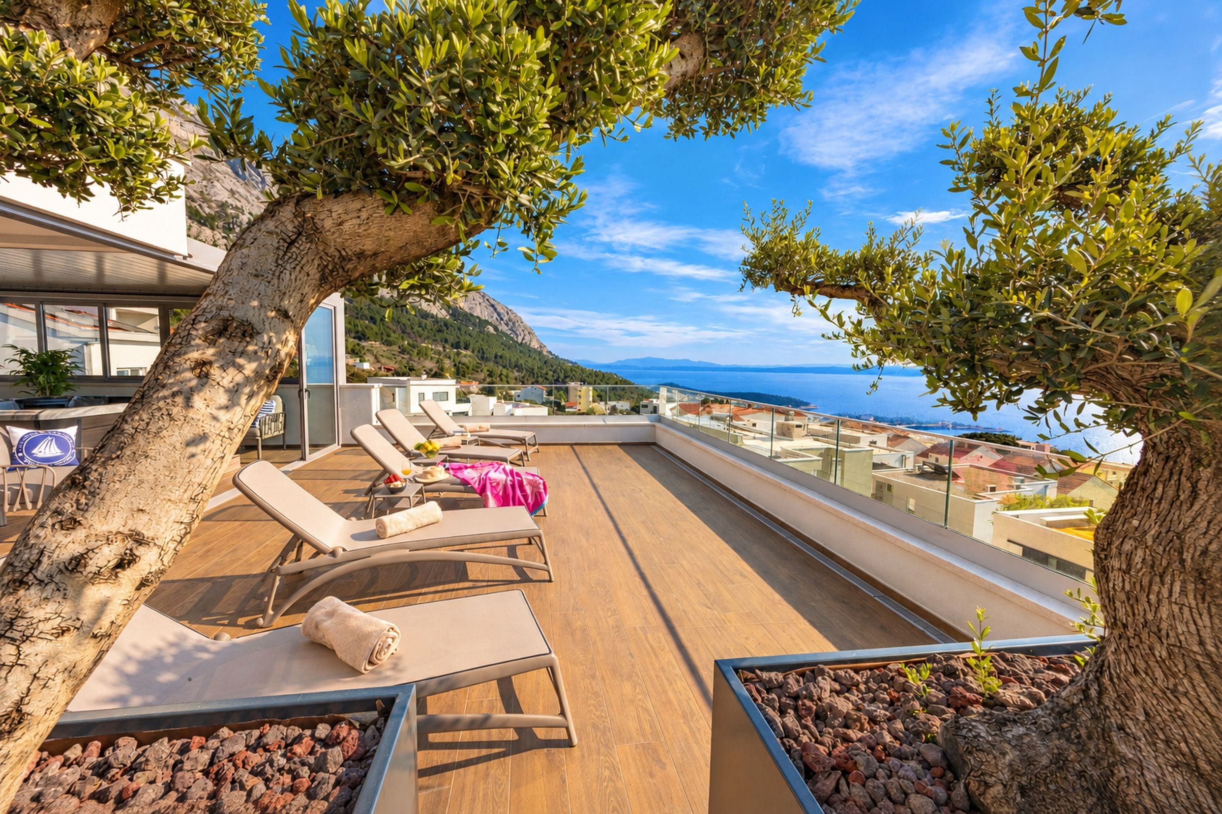 Eine Dachterrasse mit Liegestühlen, einem Baum und Blick auf das Meer und die Gebäude.