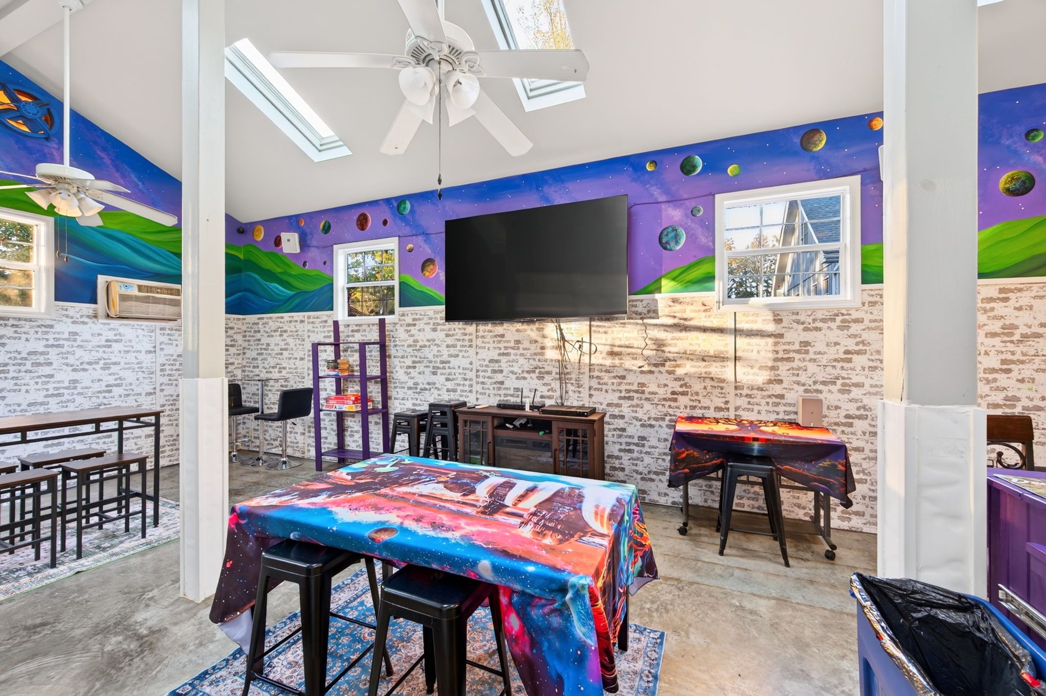 A room with a brick wall, a flat-screen TV, a table with a galaxy-themed tablecloth, and a ceiling fan.