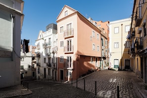 A cobblestone street with buildings on either side, including a pink building with balconies and a white building with a sign. - Graça Gate | Mouraria Ground Floor Studio (Lisbon)