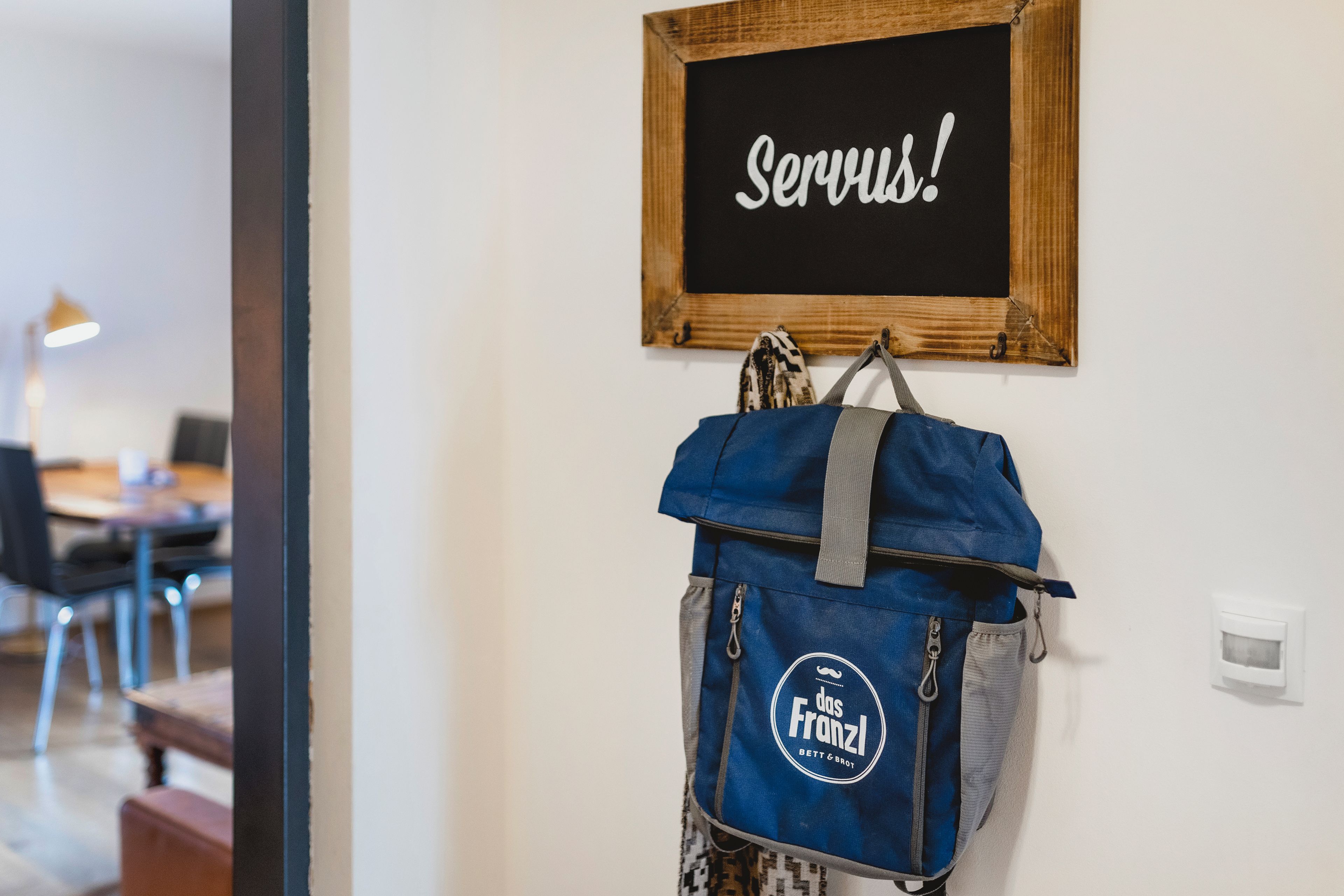 Una habitación con un cartel en la pared que dice 'Servus!' y una mochila azul colgada de un gancho.