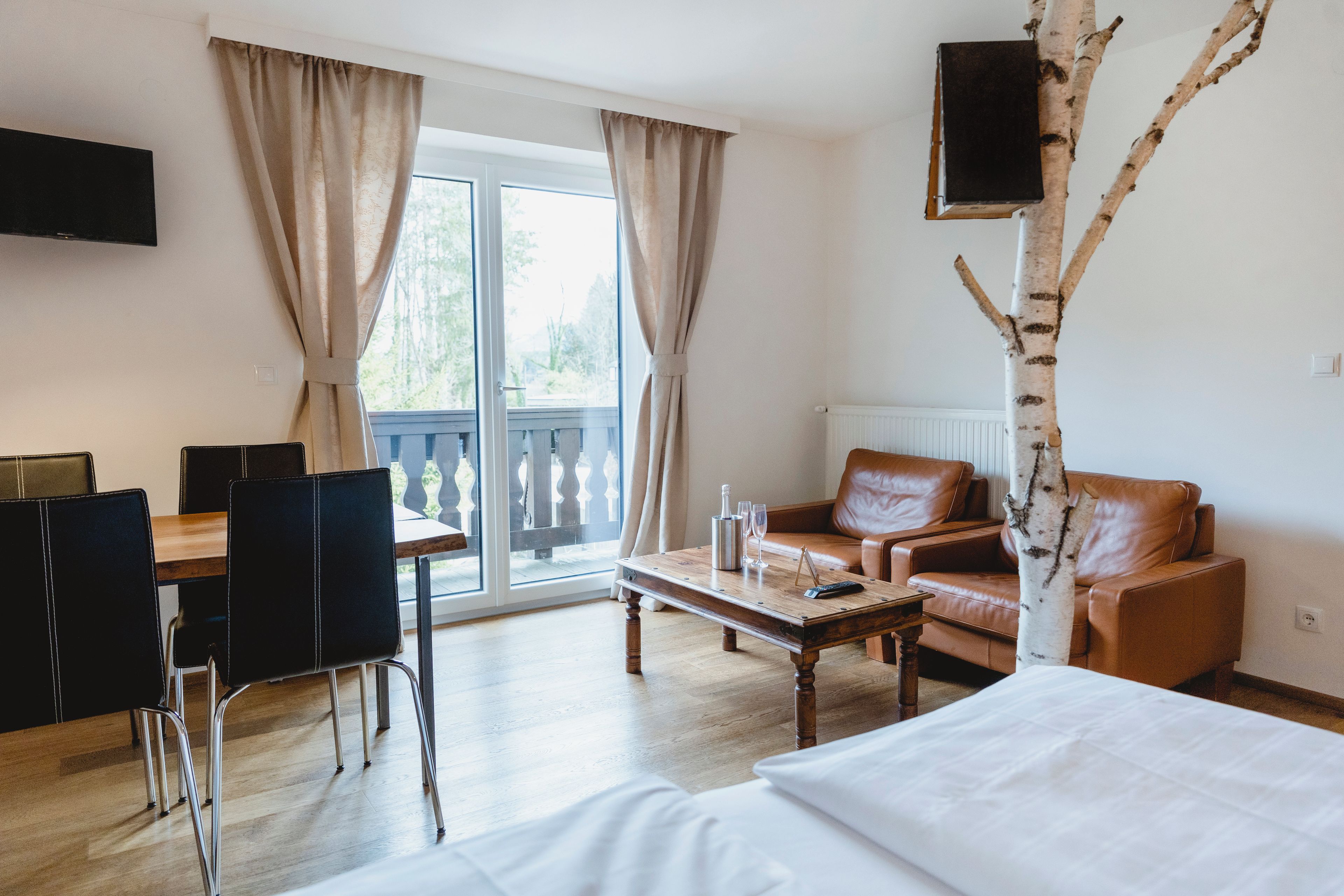 Une chambre d’hôtel comprenant un lit, un canapé, une table basse, une table à manger avec des chaises, un téléviseur fixé au mur et une grande fenêtre avec des rideaux.