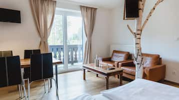 Une chambre d’hôtel comprenant un lit, un canapé, une table basse, une table à manger avec des chaises, un téléviseur fixé au mur et une grande fenêtre avec des rideaux.