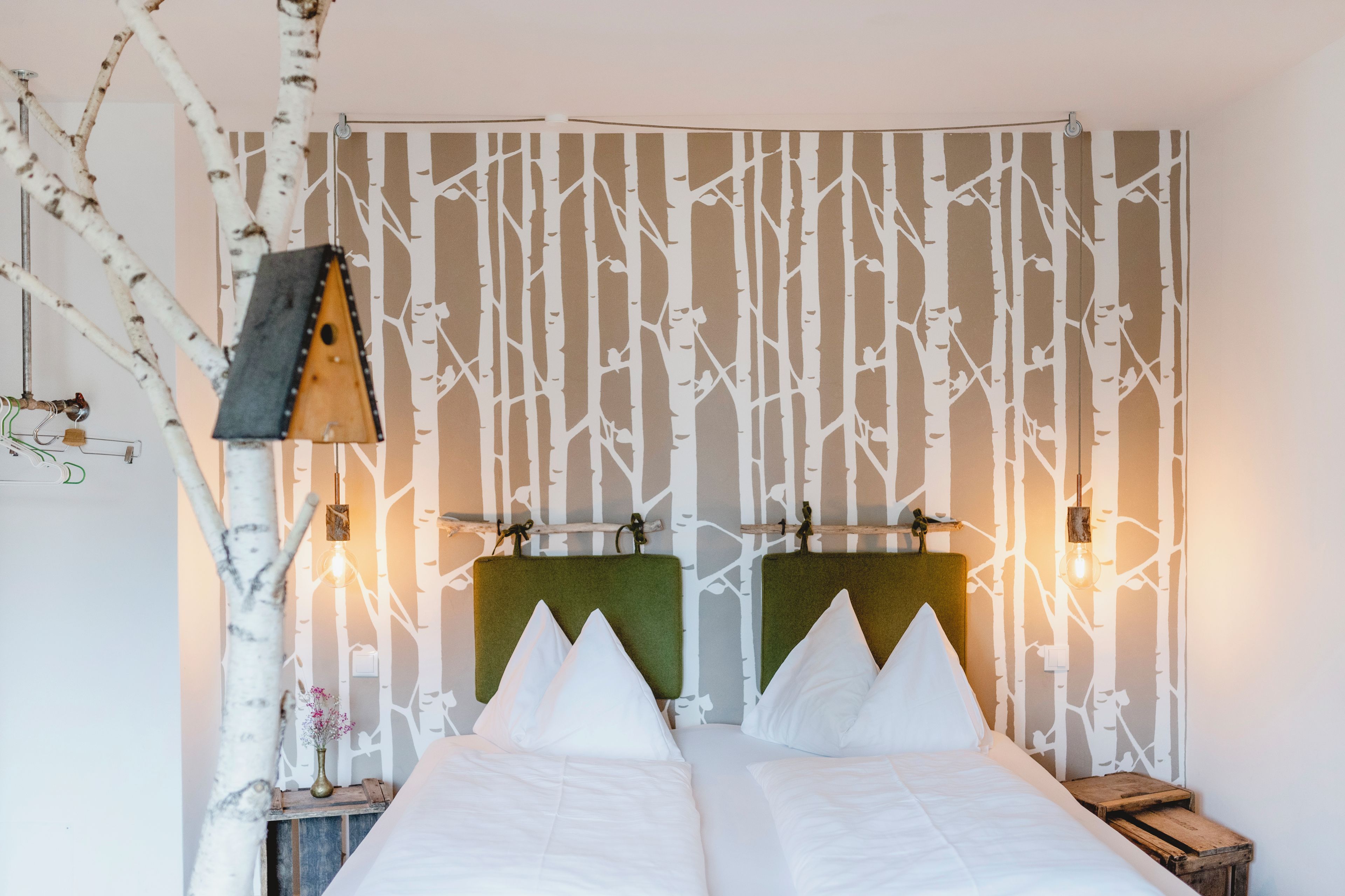 Una cama doble con cabecera, una mesita de noche con lámpara, y una pared con diseño de rama de árbol.