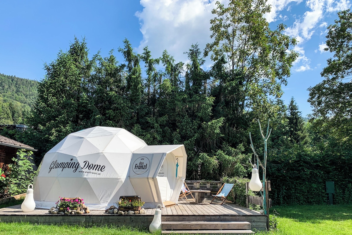 Una estructura en forma de cúpula con la leyenda 'Clamping Dome' y una dirección web, rodeada por una terraza de madera, mobiliario exterior y un área con césped.