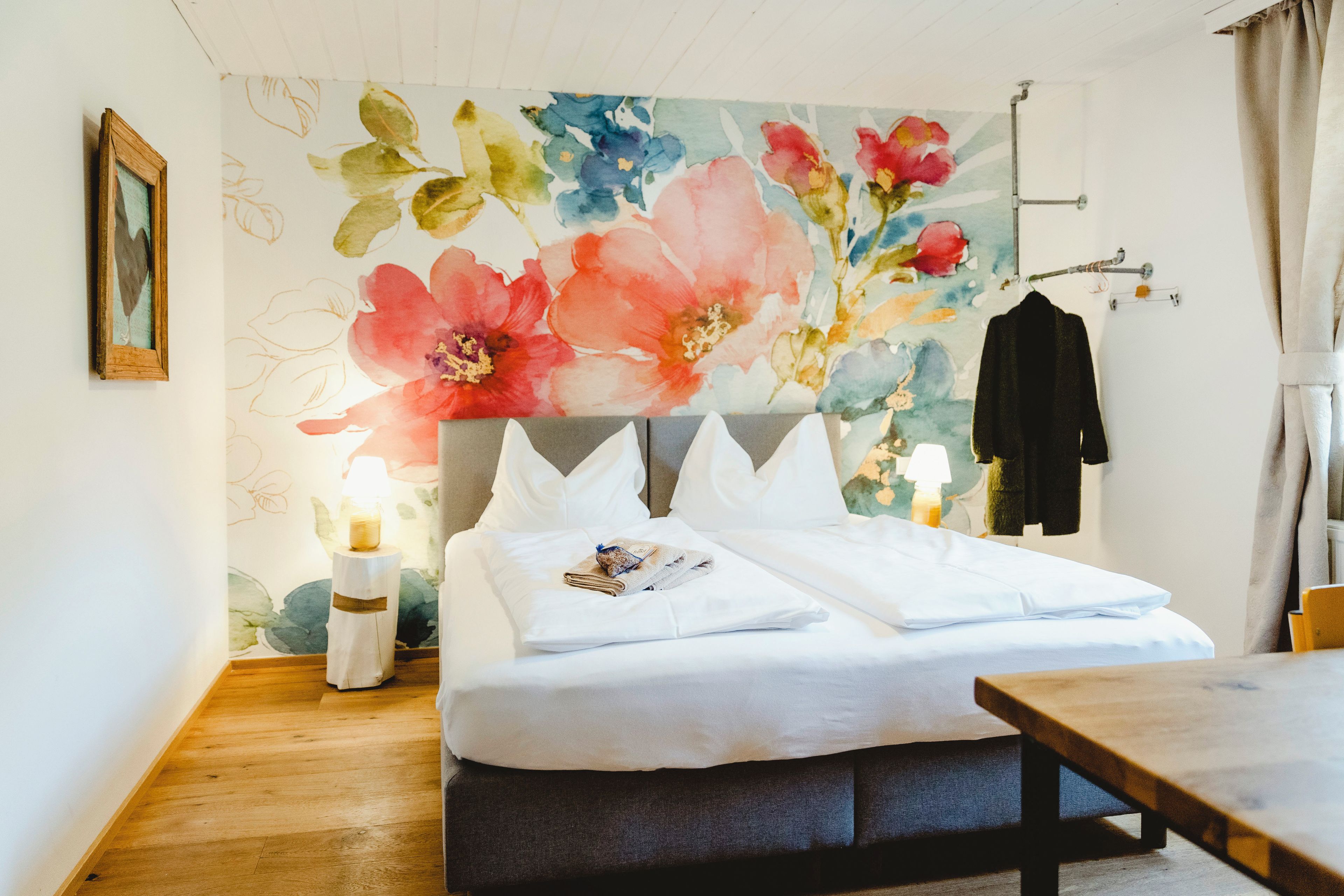 Un dormitorio con cama, una mesita de noche de madera, una lámpara, un cuadro en la pared y un perchero.