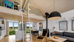 A modern interior with a loft bedroom, dining area, and living space, featuring wooden floors, a black lamp, and artwork. - 4 Star Holiday Home in Lokken (Lokken)