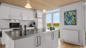 A modern kitchen with white cabinetry, a central island, stainless steel appliances, pendant lighting, and a large window offering a view of houses. - 4 Star Holiday Home in Lokken (Lokken)