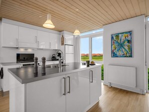 A modern kitchen with white cabinetry, a central island, stainless steel appliances, pendant lighting, and a large window offering a view of houses. - 4 Star Holiday Home in Lokken (Lokken)