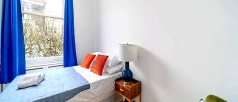 A bedroom with a bed, a nightstand, a lamp, a window with curtains, and a green chair.