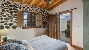 A bedroom with a stone wall, wooden ceiling, a bed with a grey bedspread, two patterned pillows, a wooden door, and a view of the outside. - Holiday Home 'La Torre' with Private Terrace, Private Garden and Wi-Fi (Santa Cruz de Tenerife)