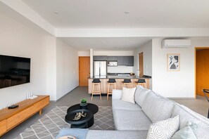 Ein modernes Wohnzimmer mit einer Couch, einem Couchtisch und einem Fernseher. Im Hintergrund ist eine Küche mit einer Bar und Hockern zu sehen.