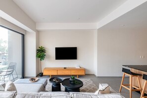 Ein modernes Wohnzimmer mit Flachbildfernseher, Holzschrank, Essbereich und großem Fenster.