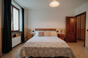 Ein Schlafzimmer mit Bett, Nachttischen, einem fensterbelaubten Fenster, einer Tür und einer Deckenleuchte.
