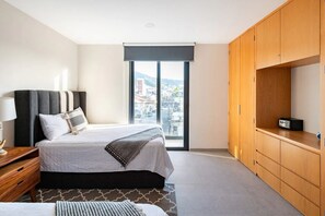 A modern hotel room with a large bed, a wooden wardrobe, a desk with a lamp, and a view of the city through a large window. - Beautiful and Central Apartment (Puerto Vallarta)