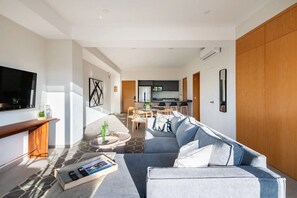 A modern living room with a grey sofa, a coffee table, and a TV. There is a kitchen in the background with a dining area. - Beautiful and Central Apartment (Puerto Vallarta)