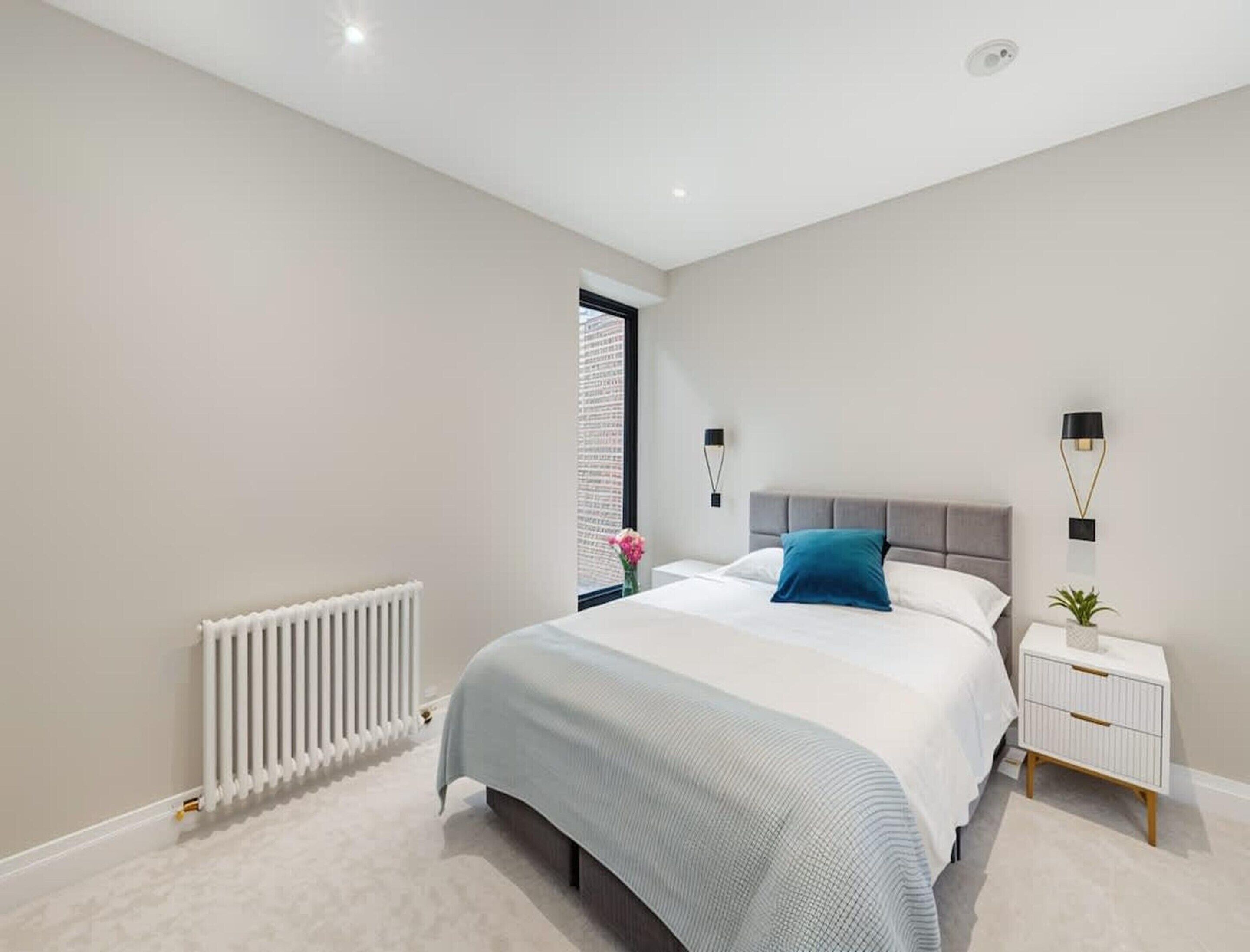 A modern bedroom with a bed, bedside tables, a radiator, and a window with blinds.