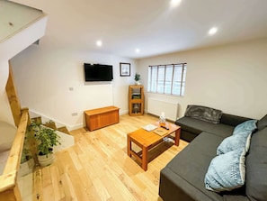A living room with a sofa, a coffee table, a wooden bench, a TV mounted on the wall, a window with blinds, and a radiator. - Broadweavers Cottage On Cotswold Way By STAE Homes (Gloucestershire)