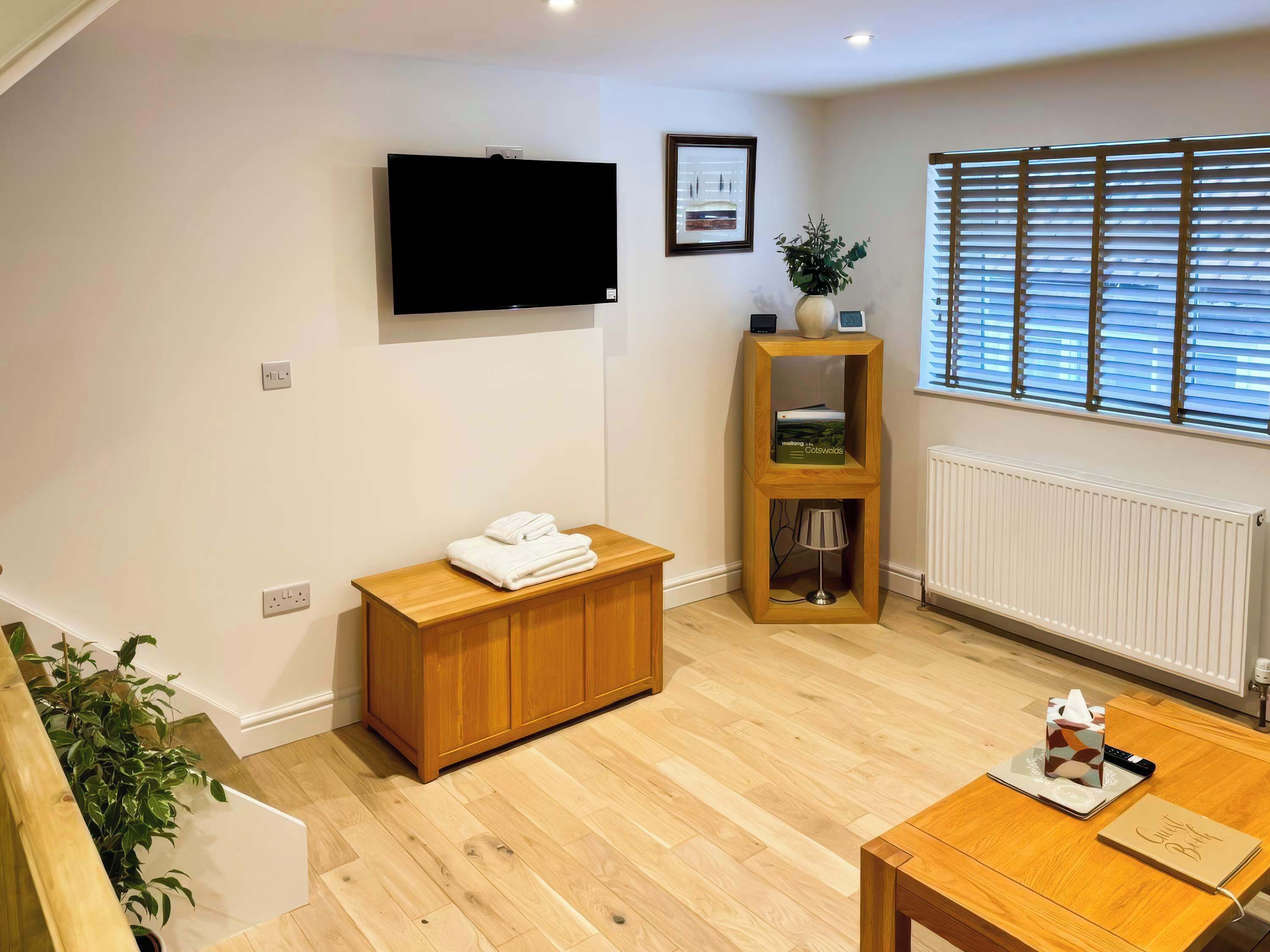 A modern living room with a flat-screen TV, wooden furniture, a radiator, and a window with blinds.