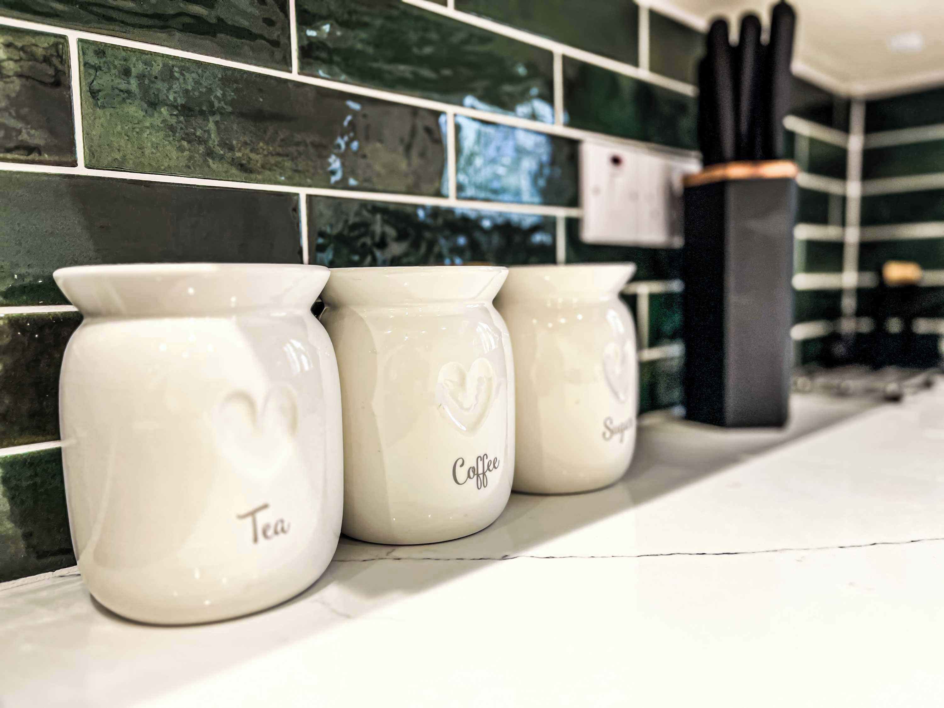 Trois récipients en céramique blanche, décorés de motifs en forme de cœur, sur lesquels sont inscrits les mots « Thé » et « Café », et qui sont posés sur un plan de travail de cuisine.