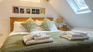 Un dormitorio con techo inclinado, un tragaluz, una cama con ropa de cama verde y blanca, una mesita de noche con una lámpara y una pared con libros enmarcados.