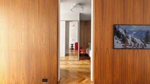 Un corridoio con pavimento in legno, con una porta che dà su una stanza con un letto, una sedia rossa e un quadro di un paesaggio montano innevato.