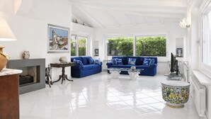 Amplia sala de estar con sofá azul, chimenea y grandes ventanales con vistas a zonas verdes.