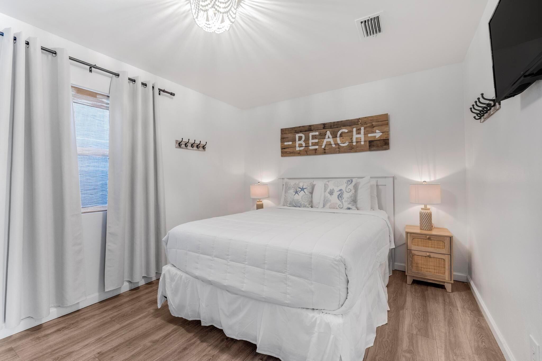 A bedroom with a bed, curtains, a bedside table, a lamp, a wall sign, and a window.