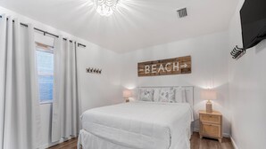 Un dormitorio con cama, cortinas, una mesita de noche, lámpara, un cartel en la pared y una ventana.