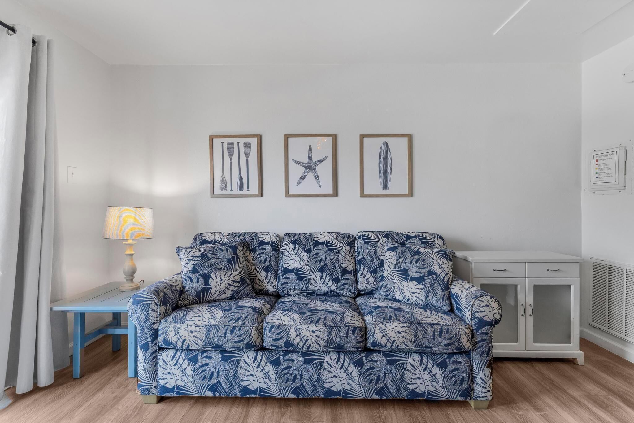 Una sala de estar con un sofá de tapicería azul y blanca, una mesita de noche con una lámpara, y tres cuadros en la pared.