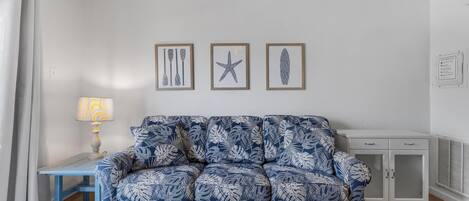 Una sala de estar con un sofá de tapicería azul y blanca, una mesita de noche con una lámpara, y tres cuadros en la pared.