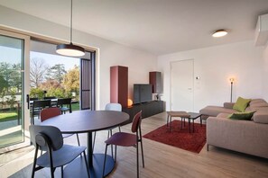 Un salon moderne comprenant un coin repas, un canapé, un téléviseur et une grande fenêtre donnant sur la verdure.