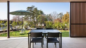Eine Terrasse im Freien mit Tisch und Stühlen, einem grünen Sonnenschirm und Blick auf Bäume und ein Gebäude im Hintergrund.