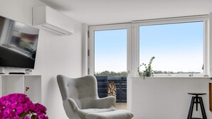 A modern living room with a grey armchair, a flat-screen TV, a small table, and a large window offering a view of the outdoors. - 4 Star Holiday Home in Lokken (Lokken)