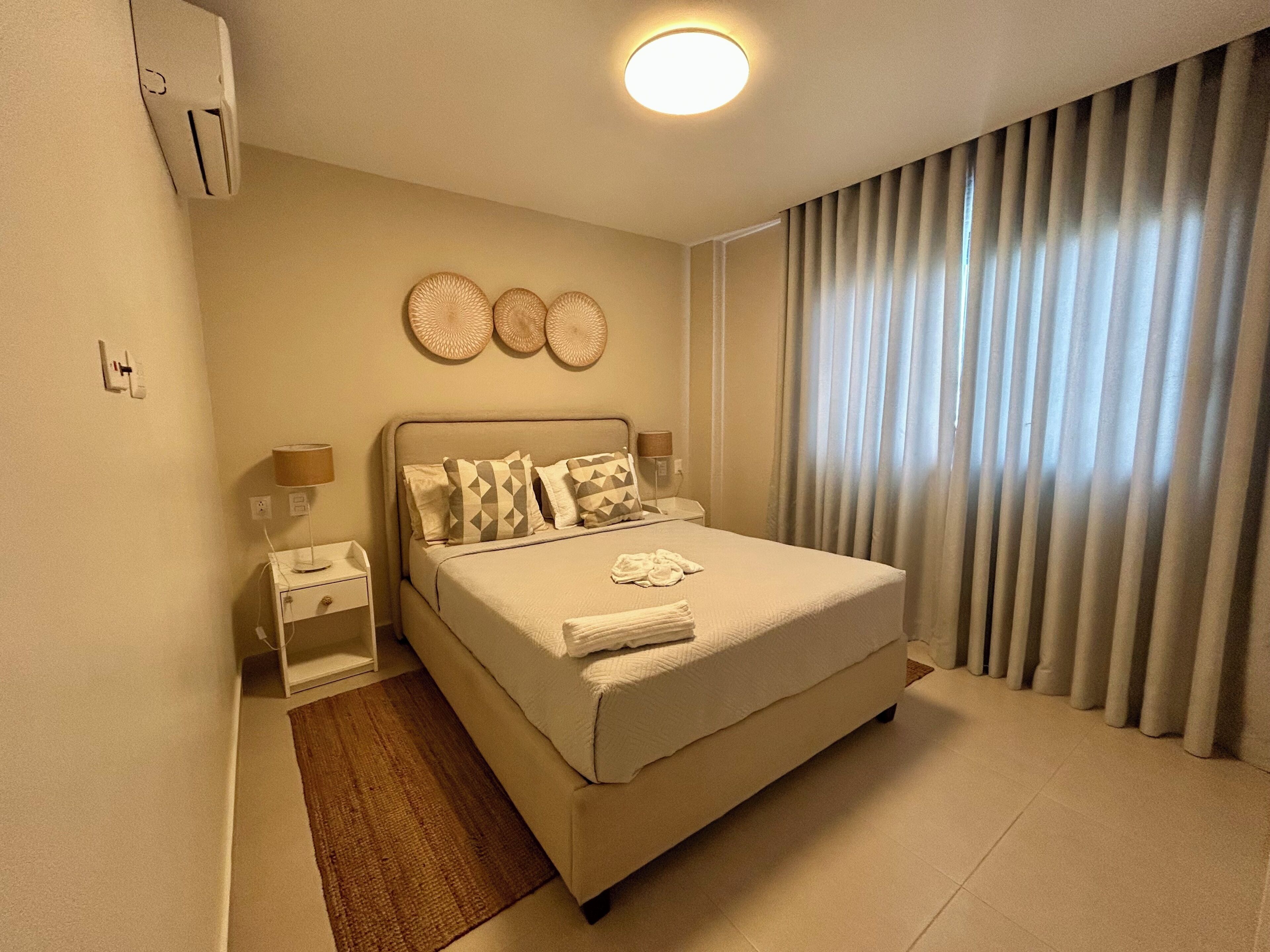 Habitación de hotel con cama, mesita de noche, lámpara, reloj en la pared y ventana con cortinas.