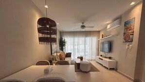 A modern living room with a dining area, a ceiling fan, a TV mounted on the wall, and a large window with curtains. - LUXURY APARTMENT IN THE BEACH (Punta Cana)