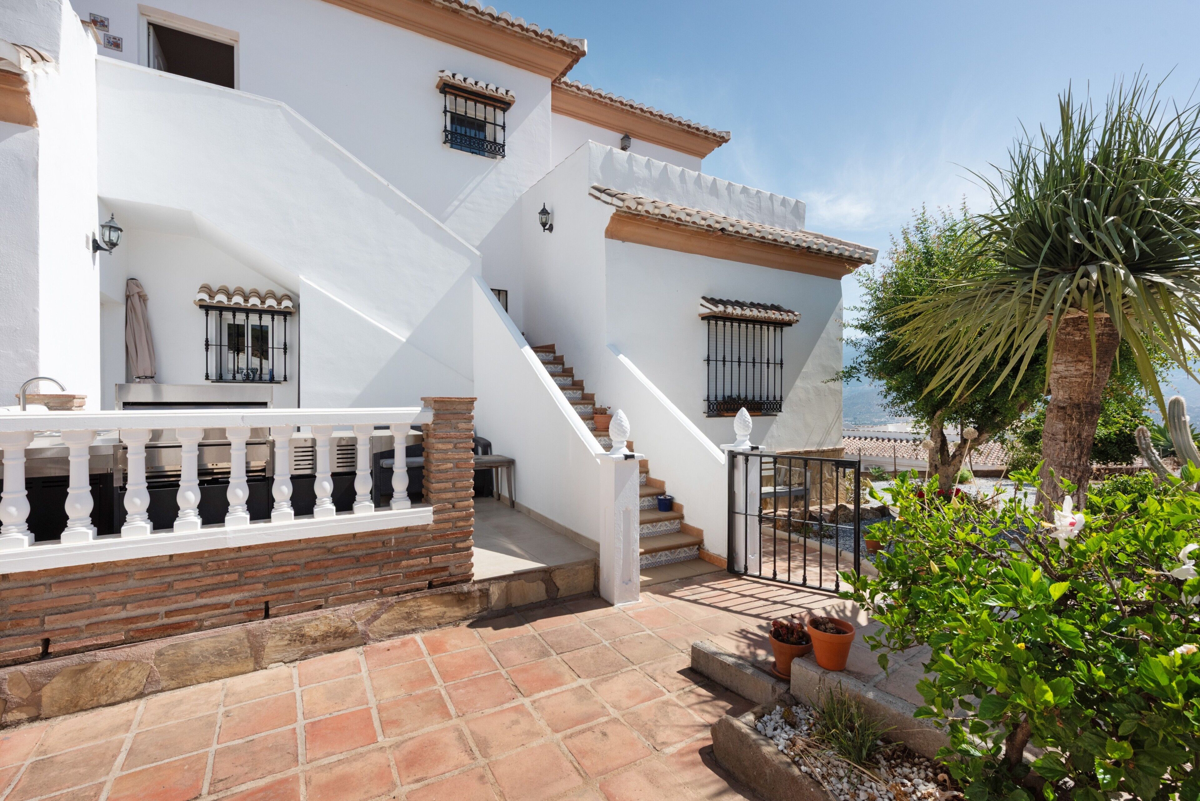 A white building with a staircase leading to a balcony, surrounded by potted plants and a palm tree.
