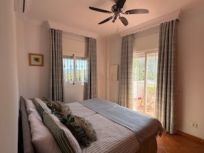 Een slaapkamer met een bed, gordijnen, een plafondventilator en een raam met uitzicht.