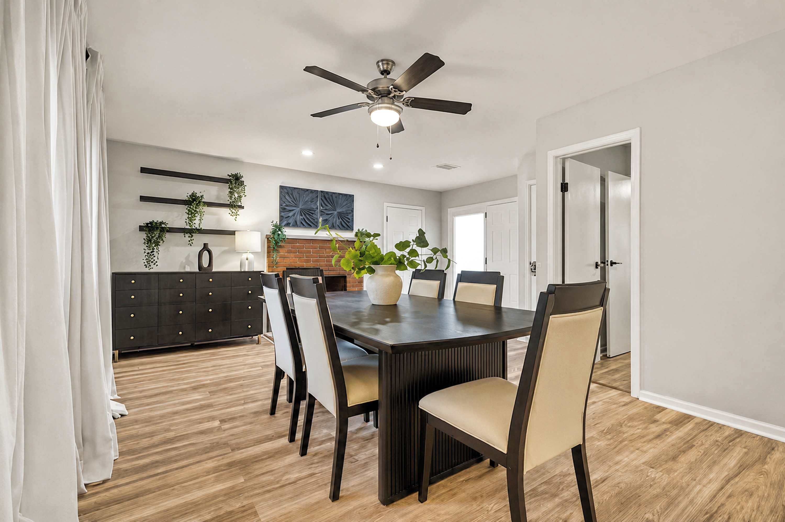 Un coin repas avec une table en bois foncé, des chaises beiges, un ventilateur de plafond et une cheminée en briques.