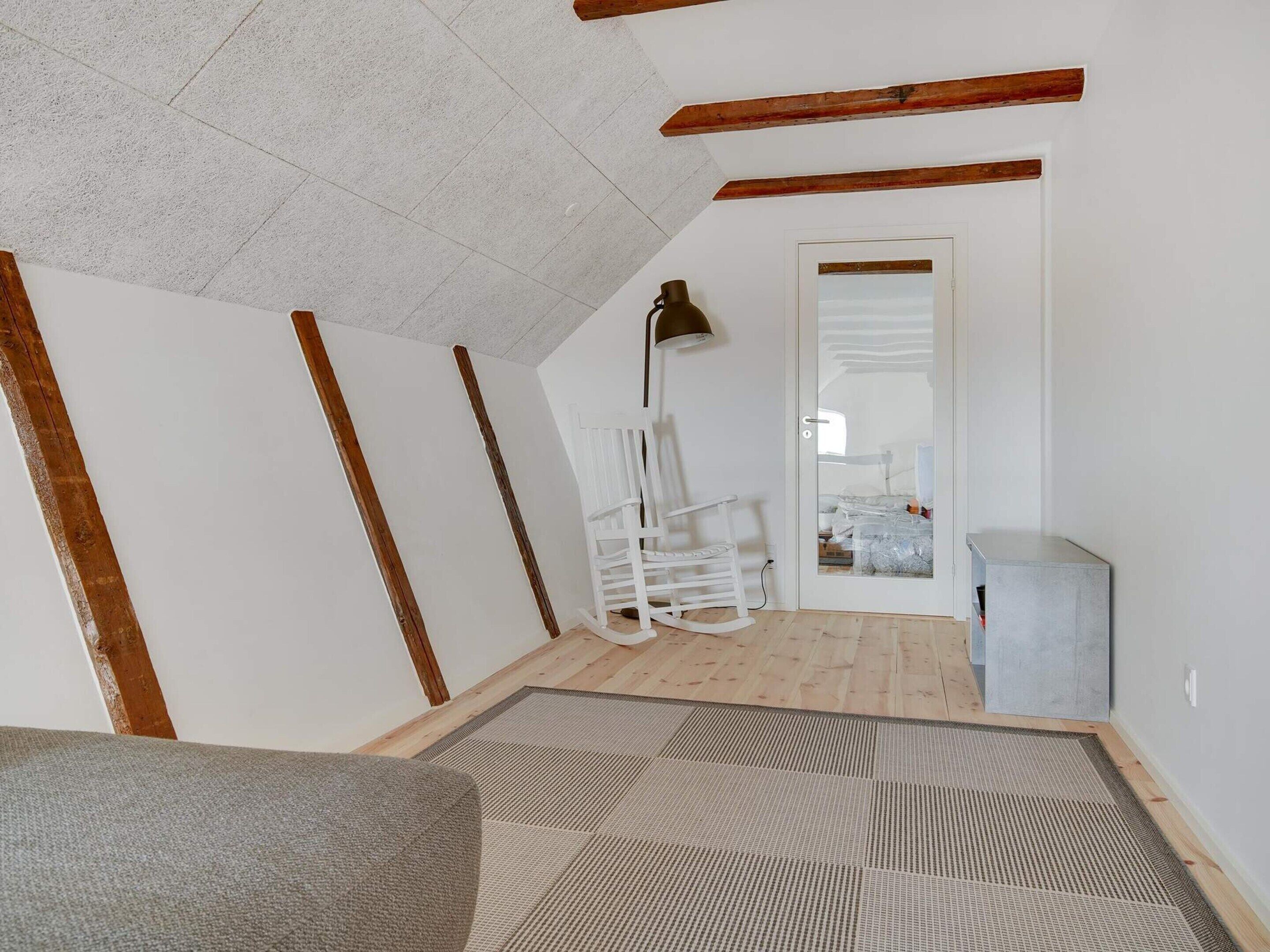 Een kamer met zichtbare houten balken, een witte muur, een schommelstoel, een lamp en een deur naar een andere kamer.
