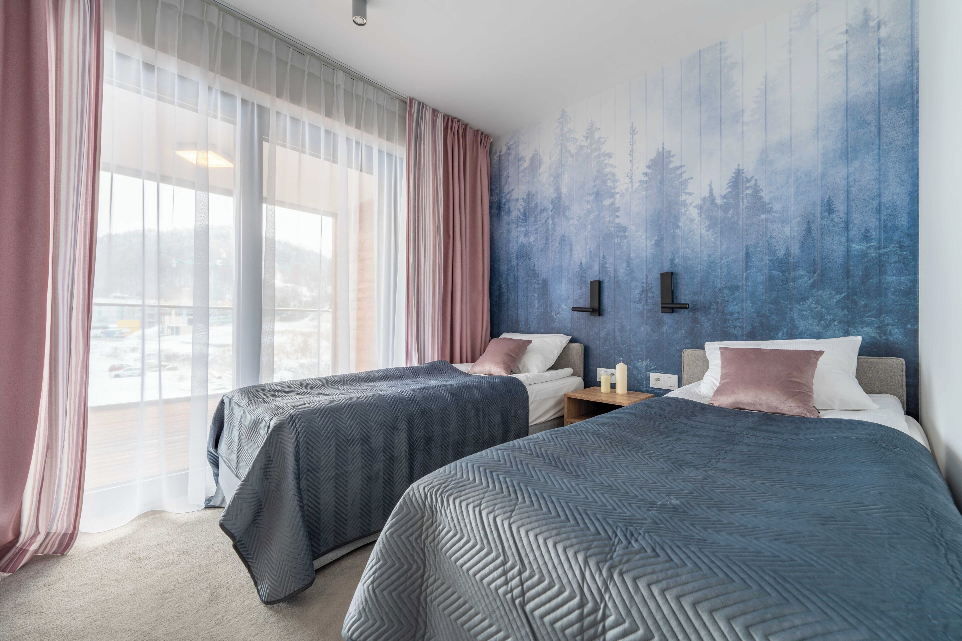 Une chambre d’hôtel avec deux lits, une grande fenêtre avec des rideaux légers, une fresque murale et une table de chevet avec une lampe.