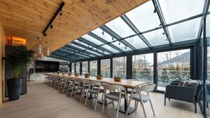A modern interior with a long dining table, chairs, a sofa, and a large window offering a view of the city.