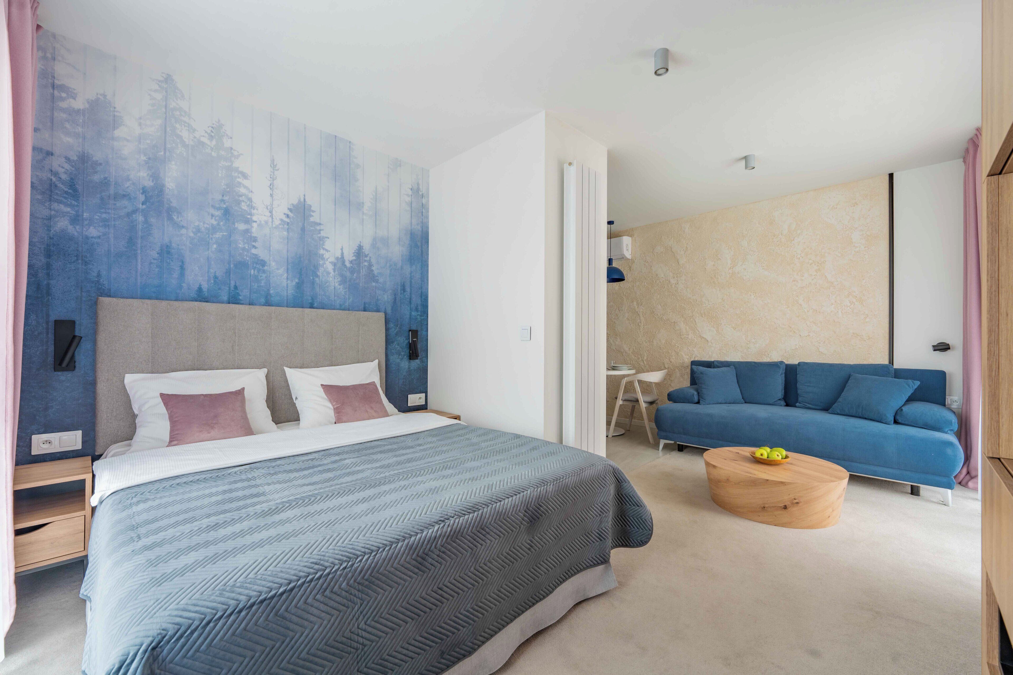 A bedroom with a bed, a blue sofa, a round wooden coffee table, and a wall with a forest mural.