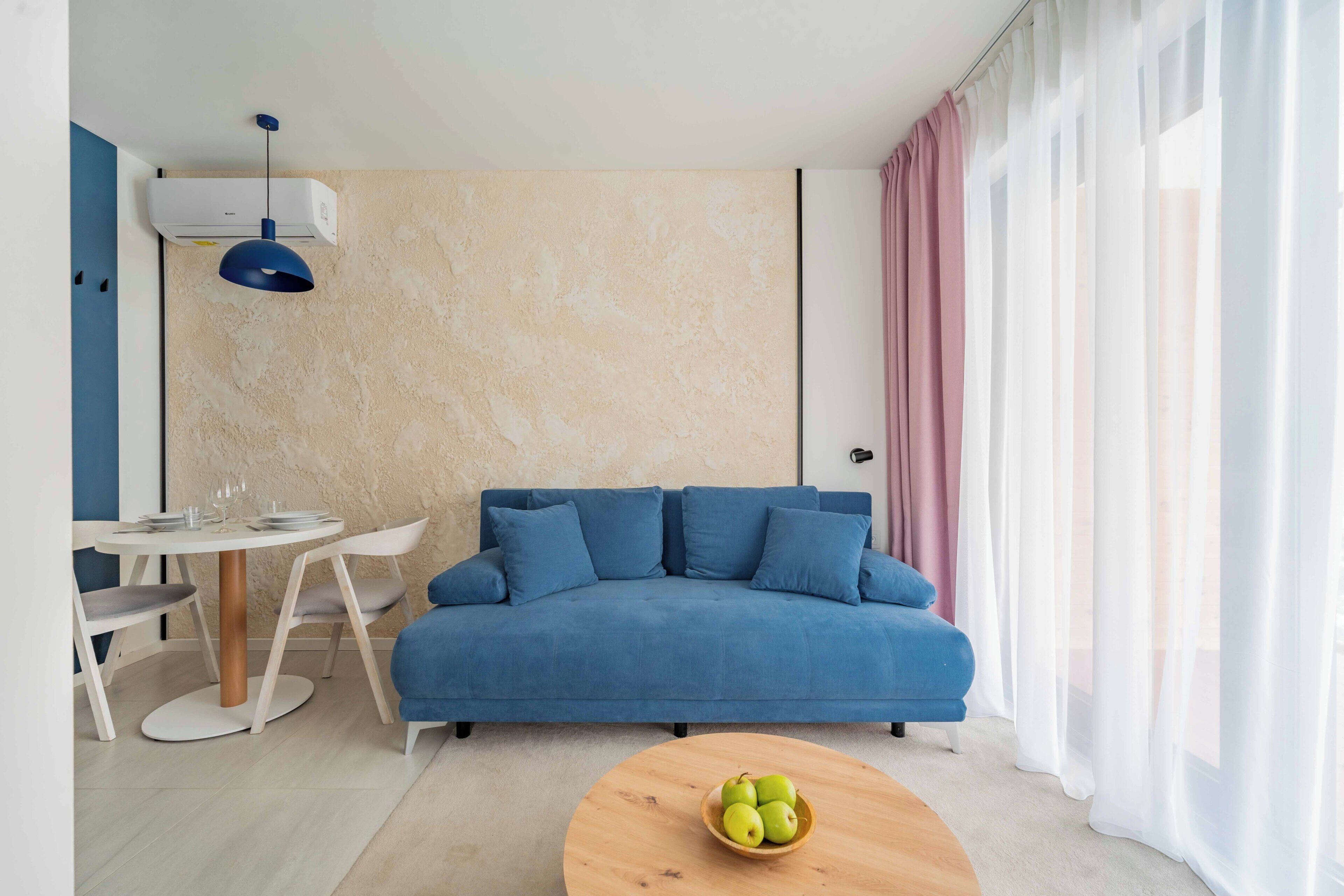 A modern living room with a blue sofa, a round wooden coffee table with apples, a white dining table with chairs, and a pendant light.