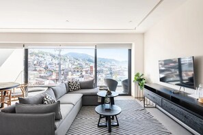 Ein modernes Wohnzimmer mit Sofa, Couchtisch und Fernseher, mit Blick auf die Stadt durch große Fenster.