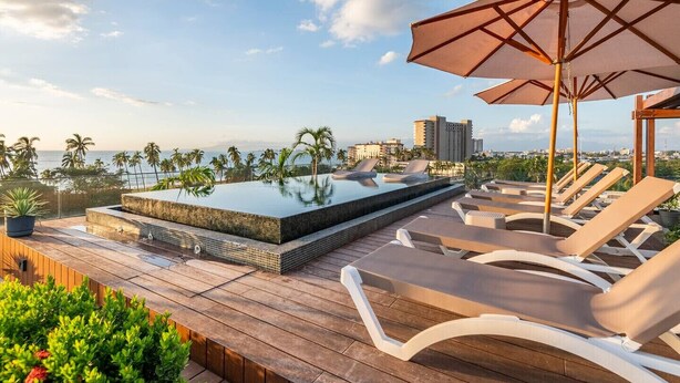 A rooftop infinity pool with lounge chairs and umbrellas, surrounded by greenery and overlooking a cityscape. - 402 (Puerto Vallarta)