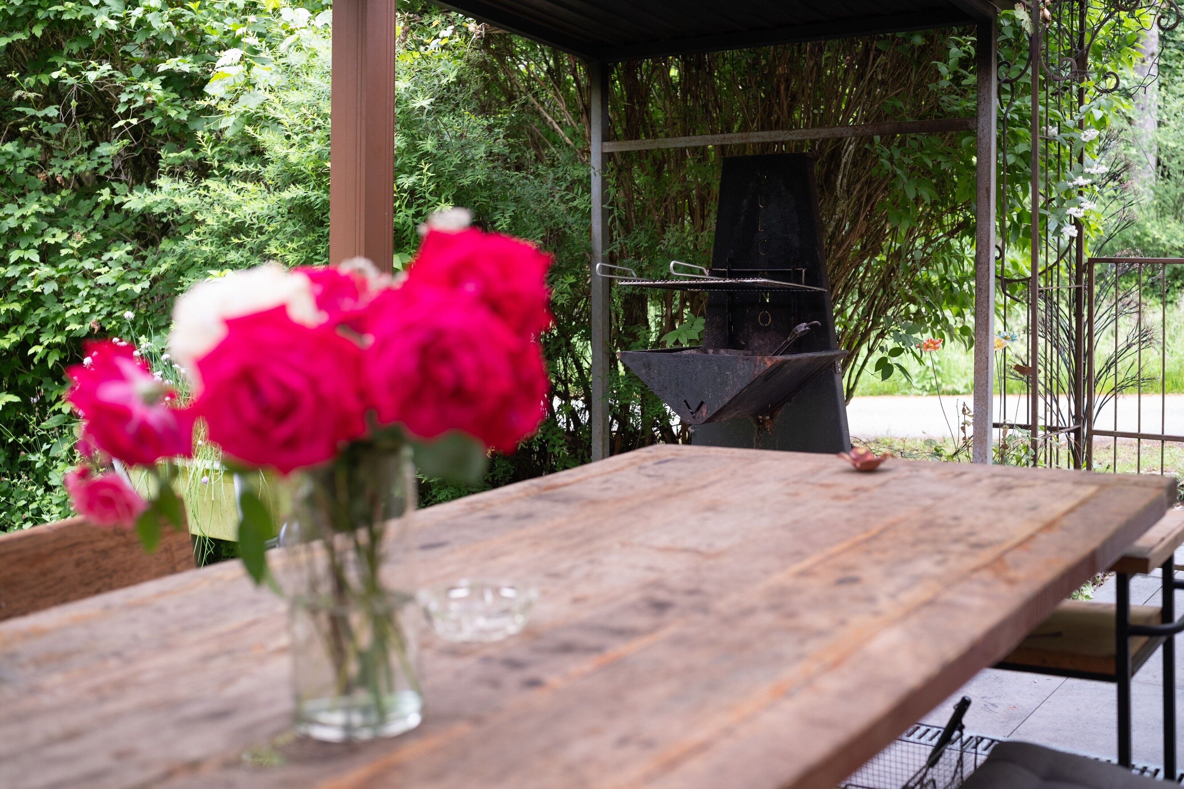A wooden table with a vase of flowers, a grill, and a metal fence in a garden setting.