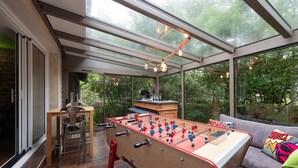 Ein raumhohes Fenster säumt einen Raum mit einem Kicker, einer Couch und einem Esstisch.